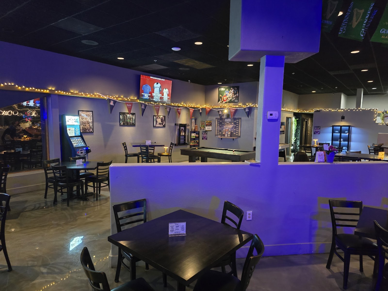 The dining room at night, lit with warm string lights, with a pool table, high-top tables and University of Arizona pennants on the wall.